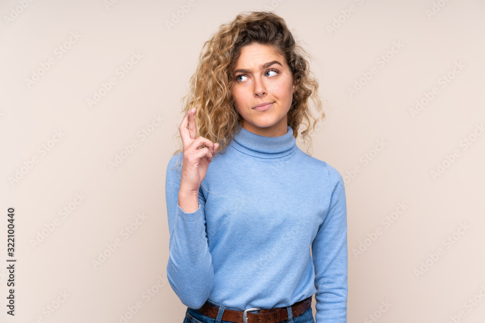 Young blonde woman with curly hair wearing a turtleneck sweater isolated on beige background with fingers crossing and wishing the best Young blonde woman with curly hair wearing a turtleneck sweater isolated on beige background with fingers crossing and wishing the best