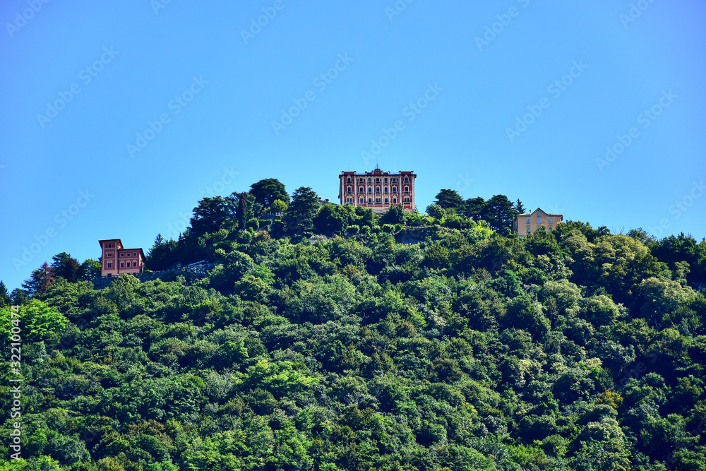 Shore of Lake Como, region Lombardia, Italy - Three white-burgundy and beige buildings on top of a green mountain with trees, blue sky in the summer afternoon.
