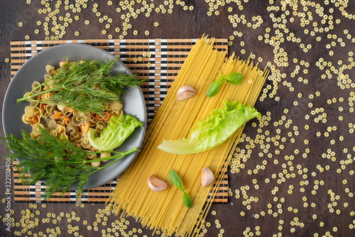 a paste of colorful shells on a gray ceramic plate is decorated with a leaf of fresh lettuce and sprigs of dill, spaghetti, baselik, lettuce and garlic on a bamboo napkin and a wooden background photo