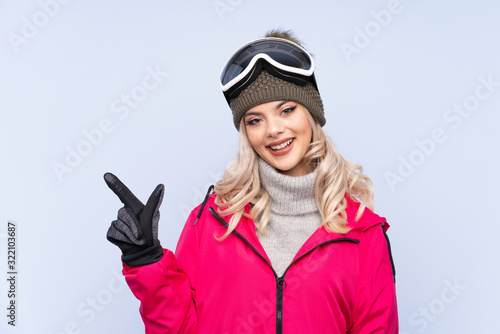 Skier teenager girl with snowboarding glasses over isolated blue background pointing finger to the side © luismolinero
