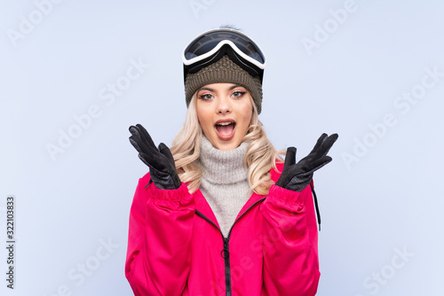 Skier teenager girl with snowboarding glasses over isolated blue background with surprise facial expression