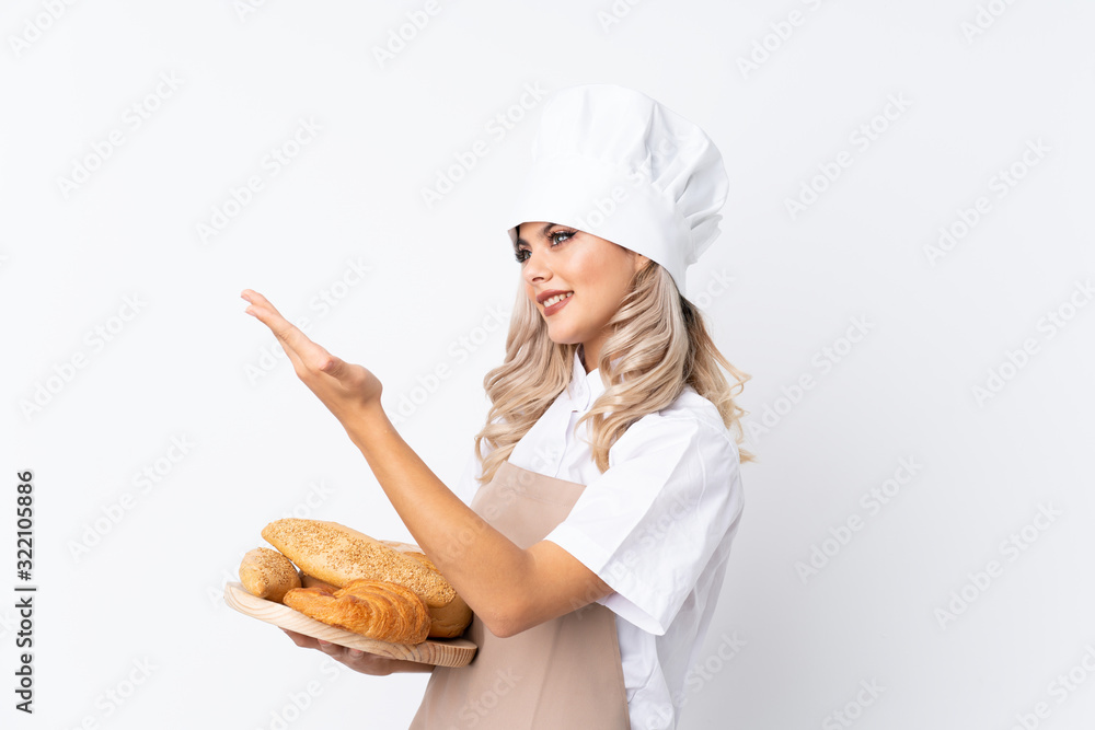 Teenager girl in chef uniform. Female baker holding a table with several breads over isolated white background extending hands to the side for inviting to come Teenager girl in chef uniform. Female baker holding a table with several breads over isolated white background extending hands to the side for inviting to come
