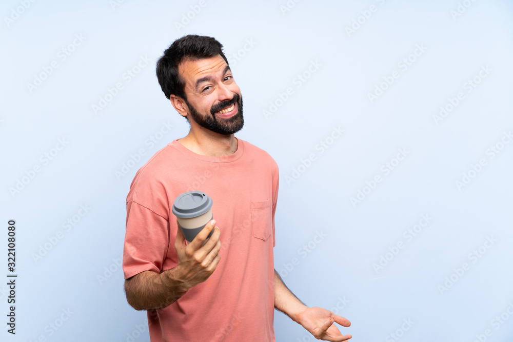 Fototapeta premium Young man with beard holding a take away coffee over isolated blue background smiling