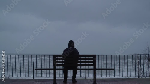 The man looks at the raging sea. A man is watching the storm. Silhouette of a man on the bench. Severe stormy sea. Big waves. Windy rainy weather. View of the dramatic ocean. The wind is blowing.
