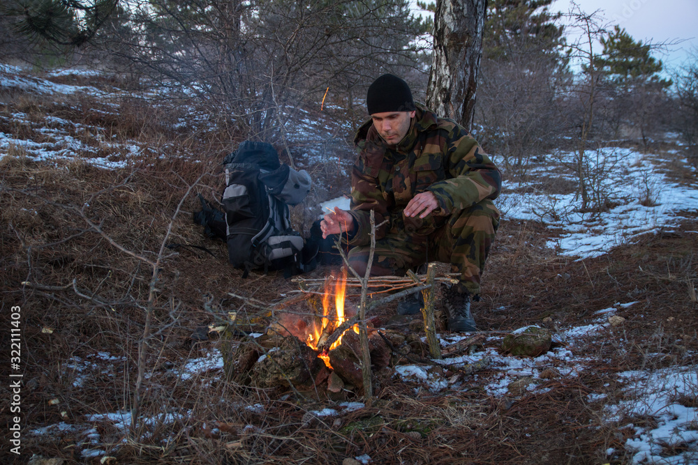 caucasian man in the camouflage sitting near the campfire in the winter forest, at night time caucasian man in the camouflage sitting near the campfire in the winter forest, at night time