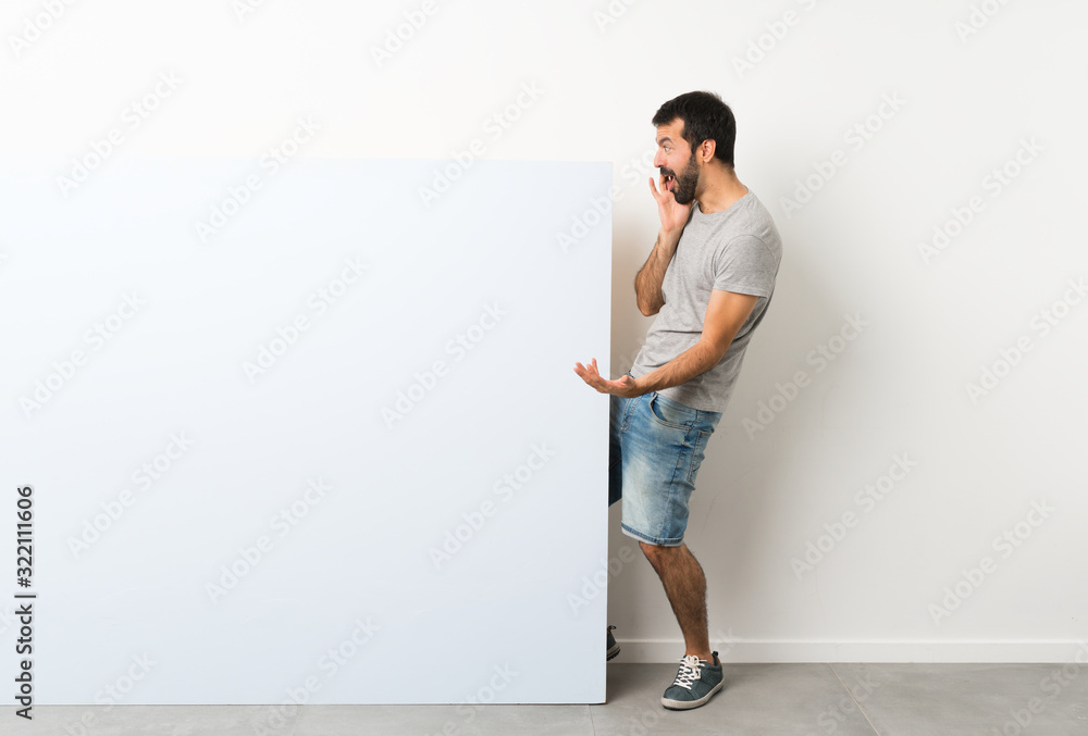 Young handsome man with beard holding a big blue empty placard with surprise and shocked facial expression Young handsome man with beard holding a big blue empty placard with surprise and shocked facial expression