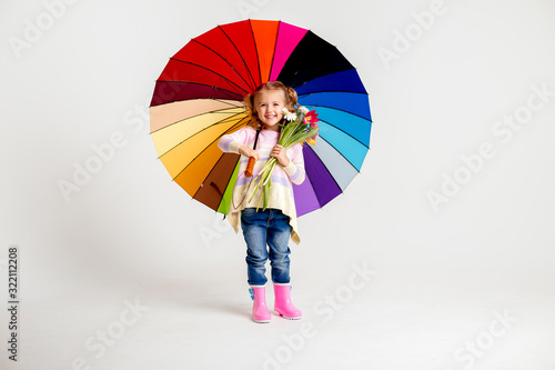 little blonde girl is smiling, standing in rubber boots holding spring flowers and a multi-colored umbrella on a white background