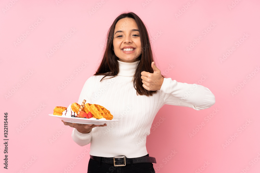 Young brunette woman holding waffles over isolated pink background showing ok sign and thumb up gesture