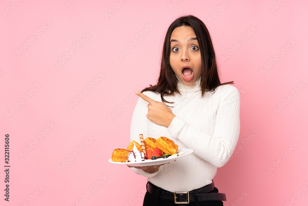 Young brunette woman holding waffles over isolated pink background surprised and pointing side Young brunette woman holding waffles over isolated pink background surprised and pointing side