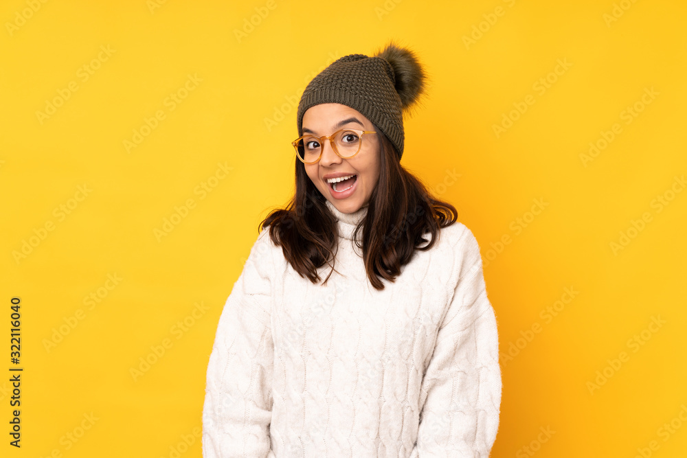 Young woman with winter hat over isolated yellow background with glasses and surprised Young woman with winter hat over isolated yellow background with glasses and surprised