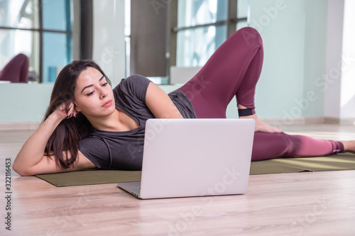 Photo of young woman practicing yoga with laptop indoor. Beautiful girl practice yoga in class. Yoga studio instructor. Blurred background.