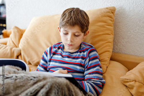 Cute blond little kid boy reading magazine or book in domestic room. Excited child reading loud, sitting on couch. Schoolkid, family, education.