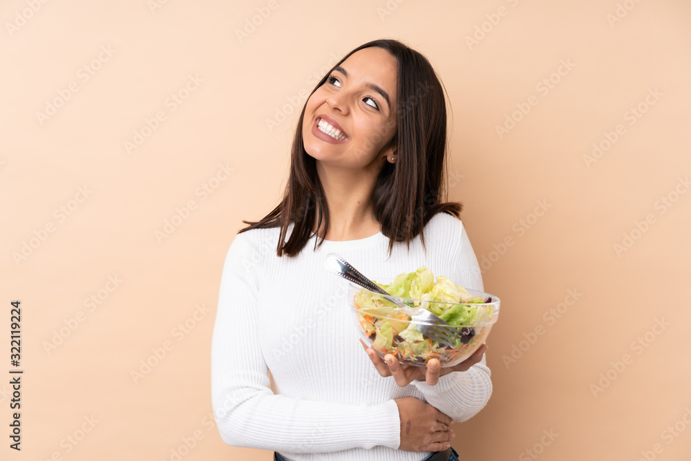 Young brunette girl holding a salad over isolated background looking up while smiling