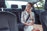 Beautiful girl secretary in business class of VIP taxi calling on phone, holding smartphone, fashionable and modern woman, cup of coffee with tea. Work on road, business meeting car. Meets passenger.