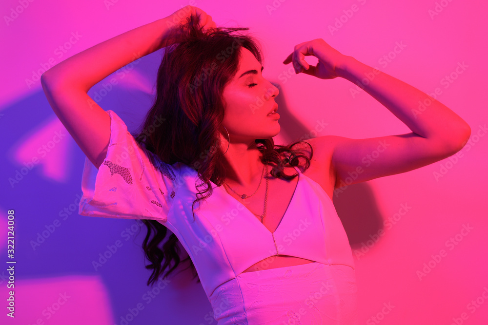 Naklejka premium Brunette with long curled hair dancing in neon. A girl with professional makeup in red and blue light posing. Sexy model in a white dress at a party.