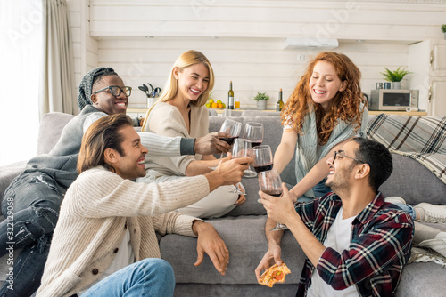 Horizontal shot of cheerful young men and women spending weekend together in cozy apartment clinking glasses with wine