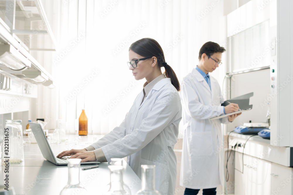 Horizontal shot of young man and woman wearing white coats standing in modern laboratory working with documents Horizontal shot of young man and woman wearing white coats standing in modern laboratory working with documents