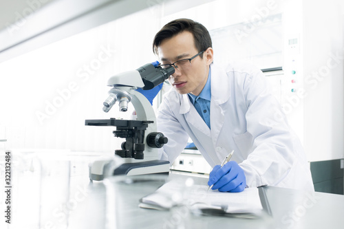 Asian medical scientist wearing white coat examining specimen using microscope, horizontal portrait