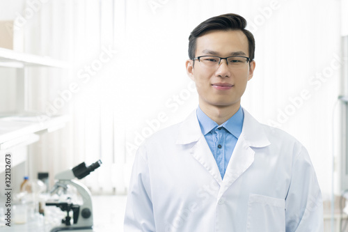 Handsome Asian medical scientist wearing white lab coat and eyeglasses looking at camera, horizontal chest up portrait