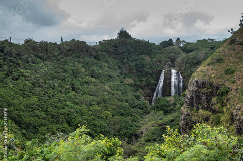 Nawiliwili, Kauai, Hawaii, USA. - January 16, 2020: White water Opaekaa Falls in wider landscape surrounded by green forest under silver sky. Brown-black rock cliffs. photo