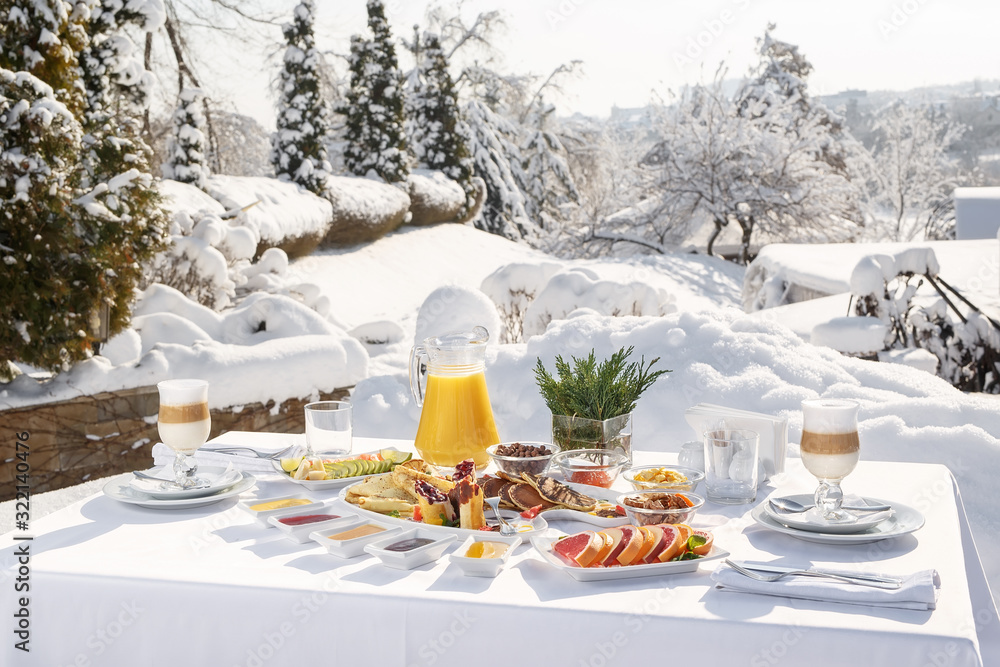 Fototapeta premium Winter Breakfast pancakes on the terrace outside the restaurant on the background of snow. Pancakes, fruit, juice, and coffee