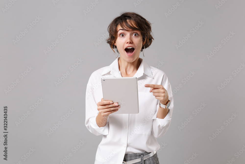Amazed young business woman in white shirt posing isolated on grey background studio portrait. Achievement career wealth business concept. Mock up copy space. Point index finger on tablet pc computer. Amazed young business woman in white shirt posing isolated on grey background studio portrait. Achievement career wealth business concept. Mock up copy space. Point index finger on tablet pc computer.