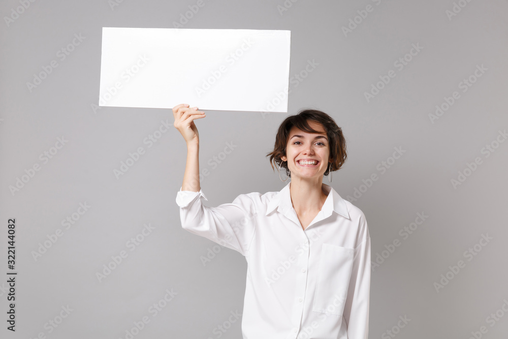 Cheerful young business woman in white shirt posing isolated on grey wall background. Achievement career wealth business concept. Mock up copy space. Hold white blank sign board with place for text. Cheerful young business woman in white shirt posing isolated on grey wall background. Achievement career wealth business concept. Mock up copy space. Hold white blank sign board with place for text.