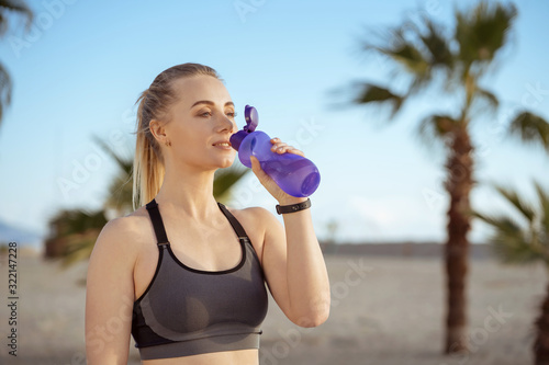 sport girl drinking water on the after training on the beach; beautiful sexy woman holding purple bottle after training;