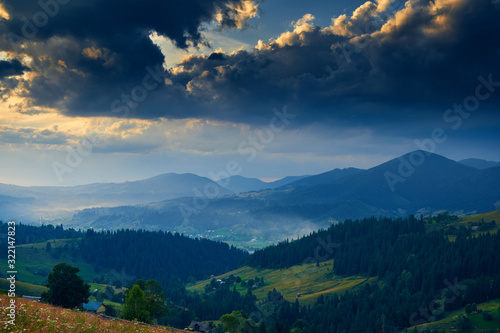 Sunset in carpathian mountains - beautiful summer landscape, spruces on hills, village, homes, dark cloudy sky and bright sun light, meadow and wildflowers