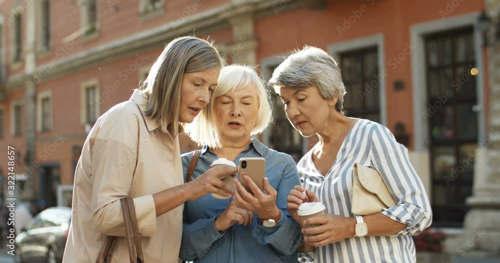 Three beautiful and cheerful Caucasian women with coffee to-go in hands laughing while scrolling photos on smartphone outdoor. Happy joyful grandmothers smiling while watching pictures on smartphone. Three beautiful and cheerful Caucasian women with coffee to-go in hands laughing while scrolling photos on smartphone outdoor. Happy joyful grandmothers smiling while watching pictures on smartphone.