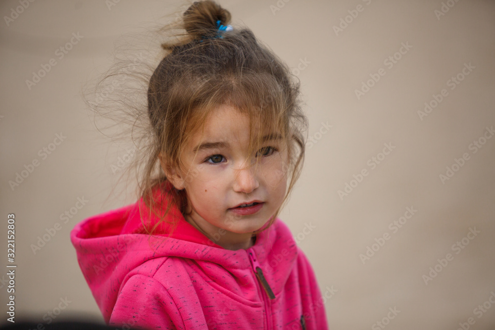 A 4-year-old girl with blonde hair in a bun with her hair flying in the wind in a pink hoodie. Girl close-up, with a blurred background, playing near the road A 4-year-old girl with blonde hair in a bun with her hair flying in the wind in a pink hoodie. Girl close-up, with a blurred background, playing near the road