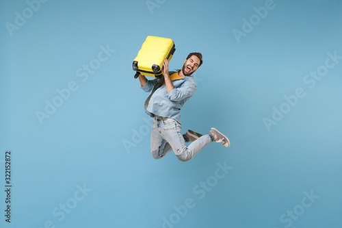 Crazy traveler tourist man in yellow casual summer clothes isolated on blue background. Male passenger traveling abroad to travel on weekend. Air flight journey concept. Jump hold suitcase screaming.