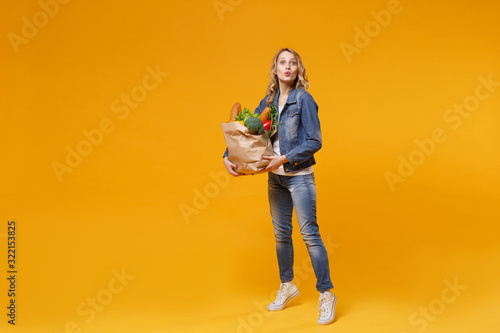 Amazed young woman in denim clothes isolated on yellow orange wall background. Delivery service from shop or restaurant concept. Hold brown craft paper bag for takeaway mock up with food products. © ViDi Studio