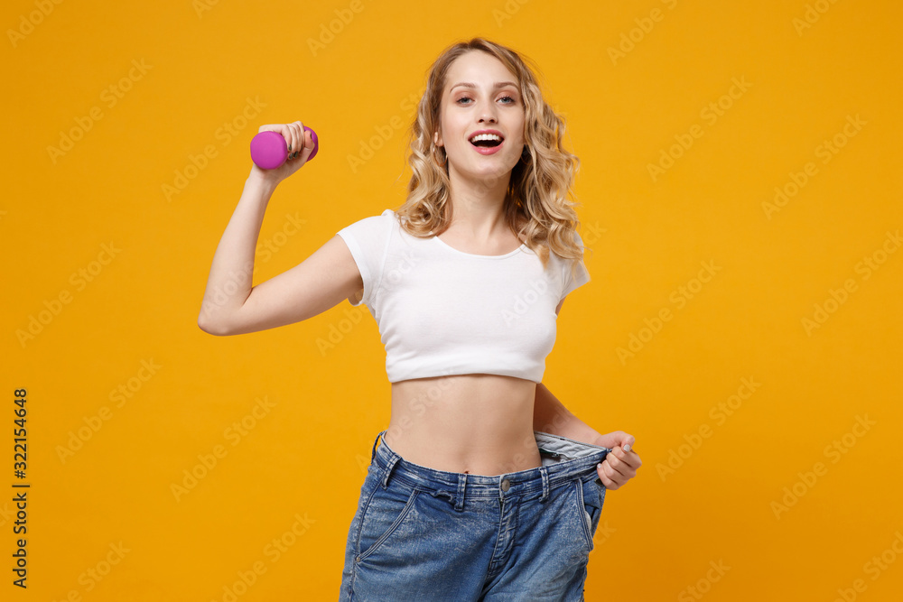 Strong young girl in white t-shirt isolated on orange background. Proper nutrition losing weight healthy sport lifestyle dieting concept. Wearing old big jeans showing her diet results hold dumbbell. Strong young girl in white t-shirt isolated on orange background. Proper nutrition losing weight healthy sport lifestyle dieting concept. Wearing old big jeans showing her diet results hold dumbbell.