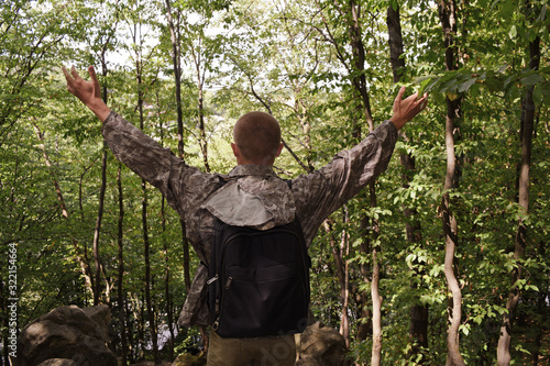 A rear view of a young man in camouflage raised his hands with a backpack.