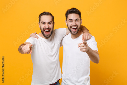 Laughing young men guys friends in white blank empty t-shirts posing isolated on yellow orange background studio portrait. People lifestyle concept. Mock up copy space. Point index fingers on camera. photo