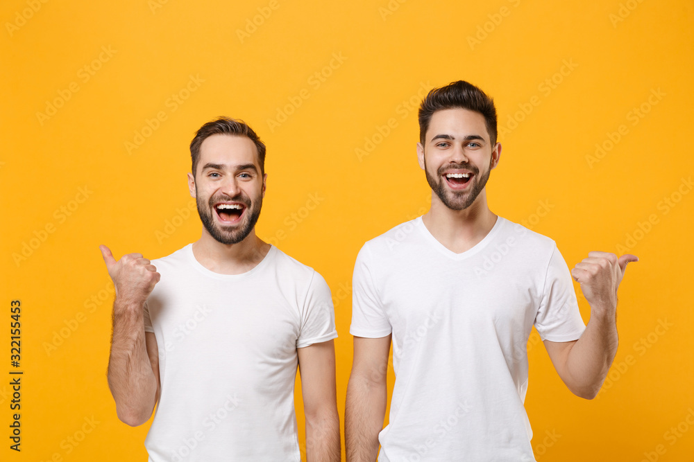 Cheerful young men guys friends in white blank empty t-shirts posing isolated on yellow orange background studio portrait. People emotions lifestyle concept. Mock up copy space. Pointing thumbs aside. Cheerful young men guys friends in white blank empty t-shirts posing isolated on yellow orange background studio portrait. People emotions lifestyle concept. Mock up copy space. Pointing thumbs aside.