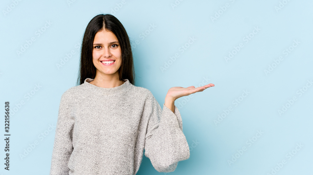 Young caucasian woman isolated on blue background showing a copy space on a palm and holding another hand on waist. Young caucasian woman isolated on blue background showing a copy space on a palm and holding another hand on waist.