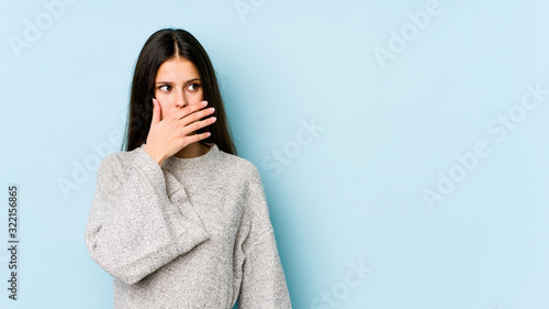 Young caucasian woman isolated on blue background thoughtful looking to a copy space covering mouth with hand. © Asier