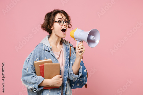 Amazed young woman student in denim clothes, eyeglasses, backpack posing isolated on pastel pink background. Education in high school university college concept. Holding books, scream in megaphone.