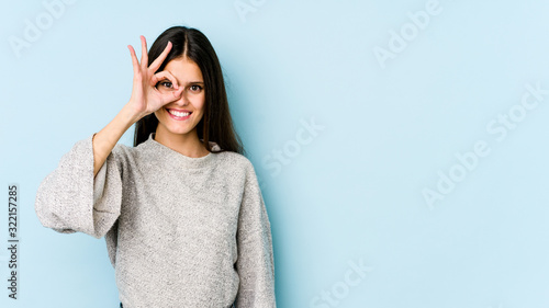 Fotografie Young caucasian woman isolated on blue background excited keeping ok gesture on eye