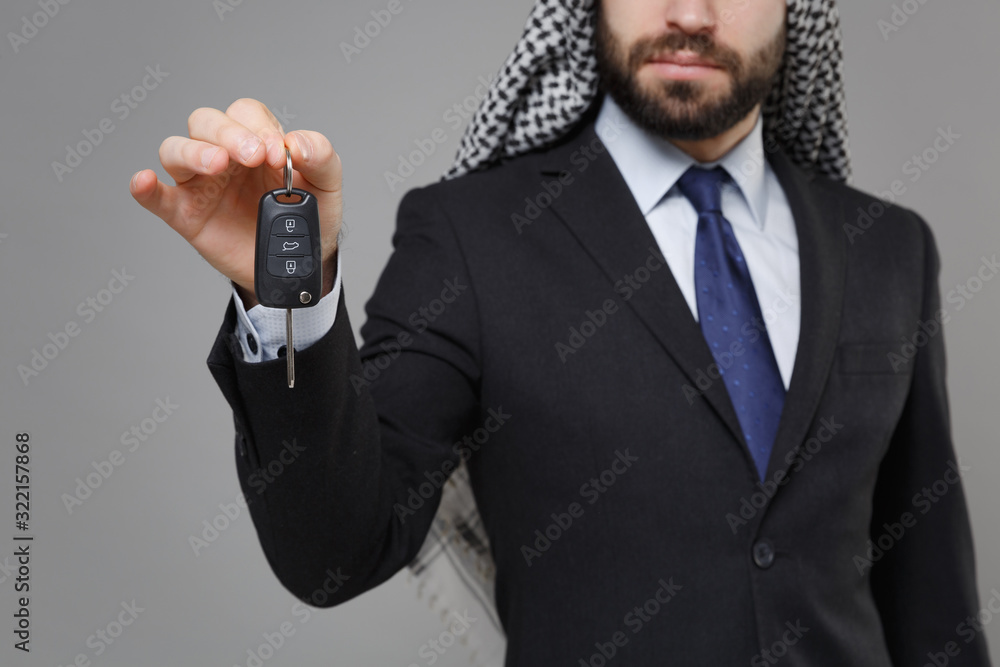 Cropped image of young bearded arabian muslim businessman in keffiyeh kafiya ring igal agal classic black suit isolated on gray background. Achievement career wealth business concept. Hold car keys. Cropped image of young bearded arabian muslim businessman in keffiyeh kafiya ring igal agal classic black suit isolated on gray background. Achievement career wealth business concept. Hold car keys.