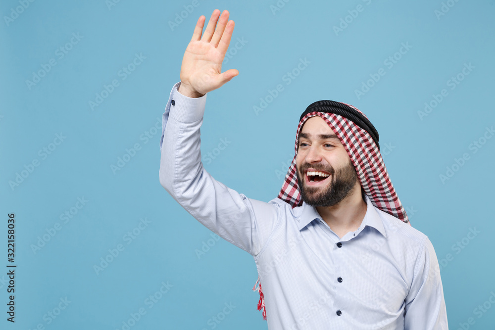 Naklejka premium Joyful bearded arabian muslim man in keffiyeh kafiya ring igal agal casual clothes isolated on pastel blue background. People religious lifestyle concept. Waving greeting with hand as notices someone.