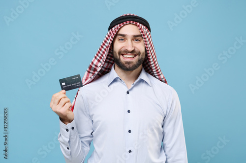 Smiling young bearded arabian muslim man in keffiyeh kafiya ring igal agal casual clothes posing isolated on pastel blue wall background. People religious lifestyle concept. Holding credit bank card. photo