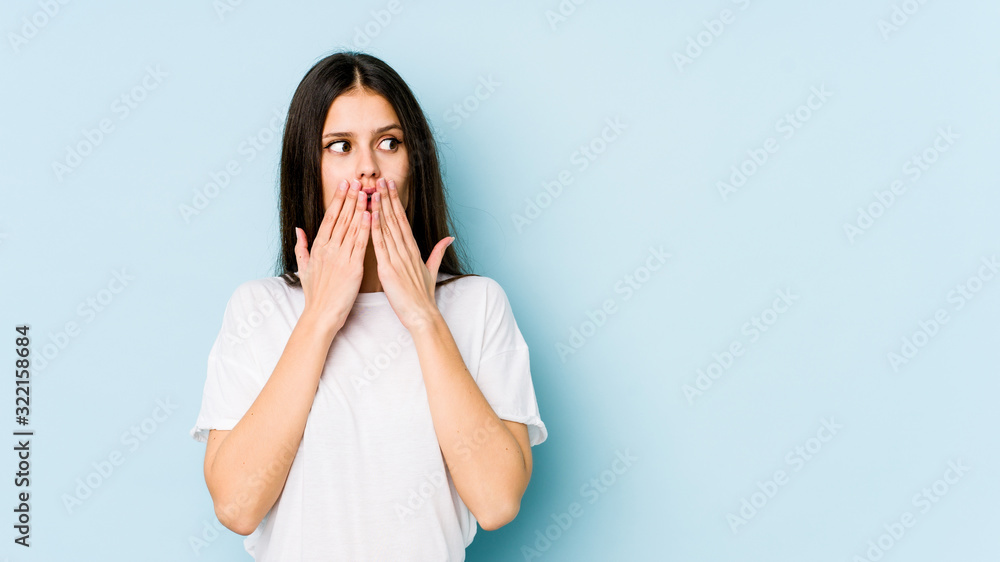 Young caucasian woman isolated on blue background thoughtful looking to a copy space covering mouth with hand. Young caucasian woman isolated on blue background thoughtful looking to a copy space covering mouth with hand.