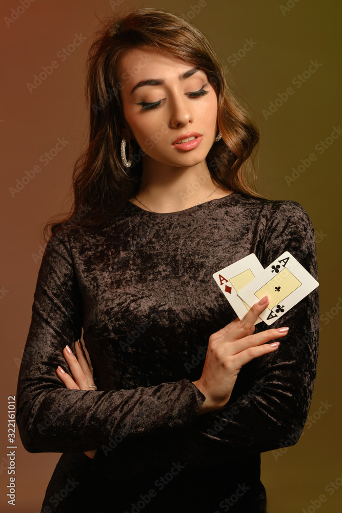 Brunette girl in black velvet dress and jewelry showing two aces, posing against colorful studio background. Gambling, poker, casino. Close-up. Brunette girl in black velvet dress and jewelry showing two aces, posing against colorful studio background. Gambling, poker, casino. Close-up.