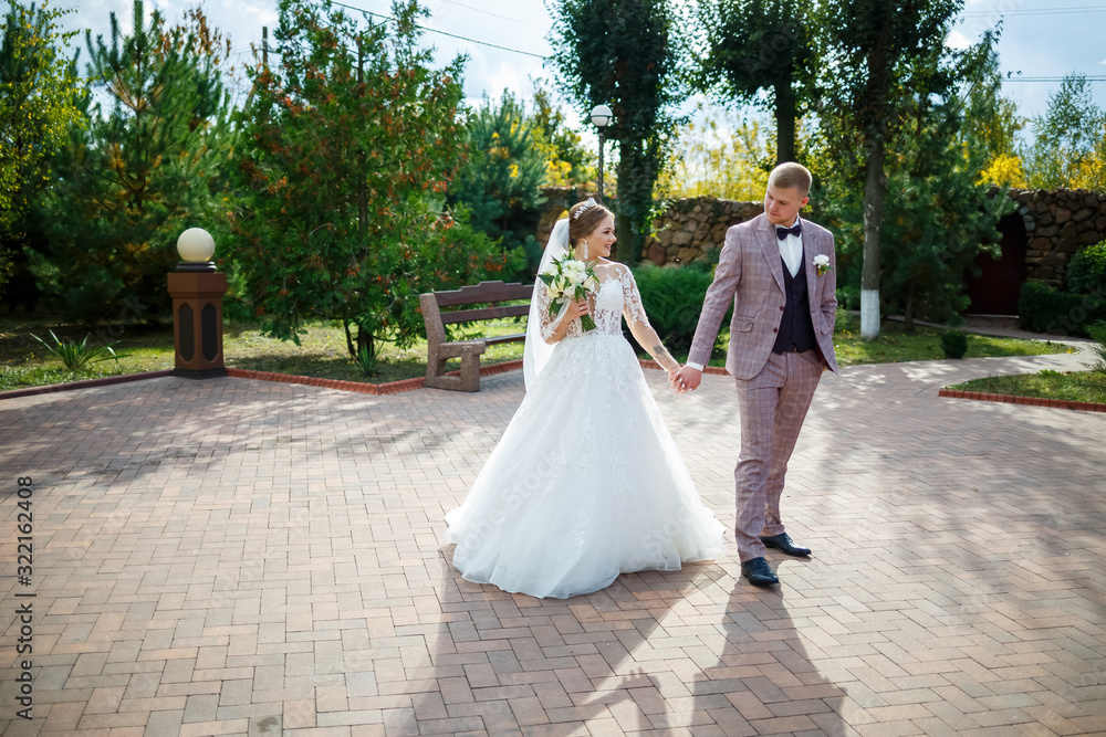 Bride in white dress and groom in costume cuddle and walk in the park