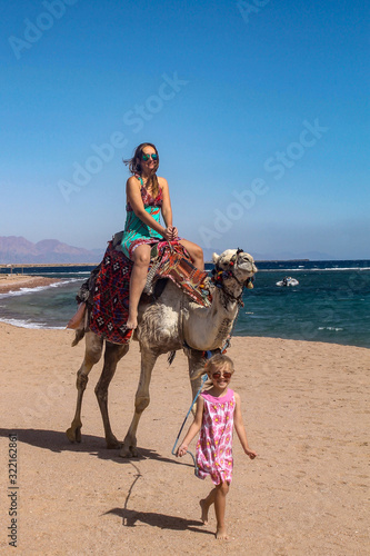 Mother and little daughter a camel, a Caucasian girl leads a camel on which her mother sits on the background of the sea and a summer beach, a child with her mother on a camel in the desert by the sea © Oleksandr
