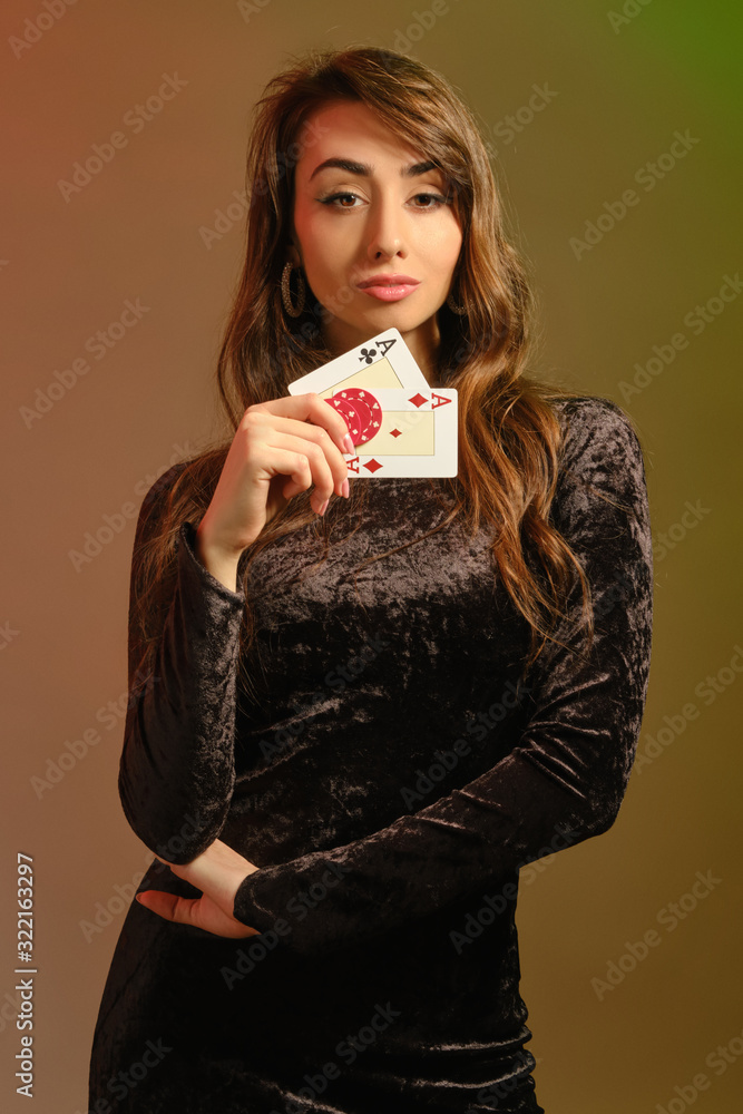 Brunette girl in black dress showing two red chips and aces, posing against colorful studio background. Gambling, poker, casino. Close-up. Brunette girl in black dress showing two red chips and aces, posing against colorful studio background. Gambling, poker, casino. Close-up.