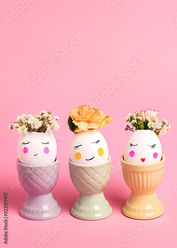Easter creative concept. Easter decorative eggs with painted cheerful faces and flowers instead of hair in the egg cups on the pink background. Copy space, close up